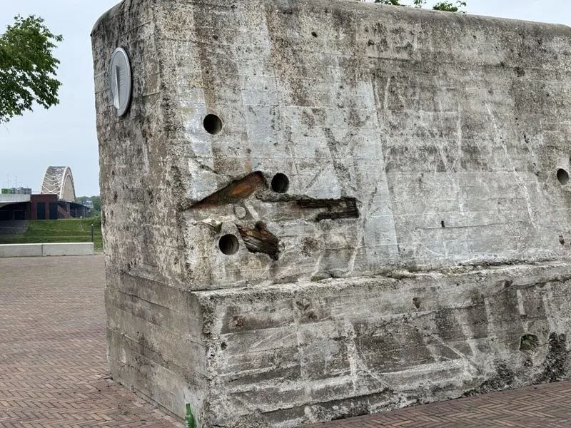 Weathered grey concrete anti-tank blocks standing on the north bank of the Waal at Nijmegen, one with traces of red-and-white paint.