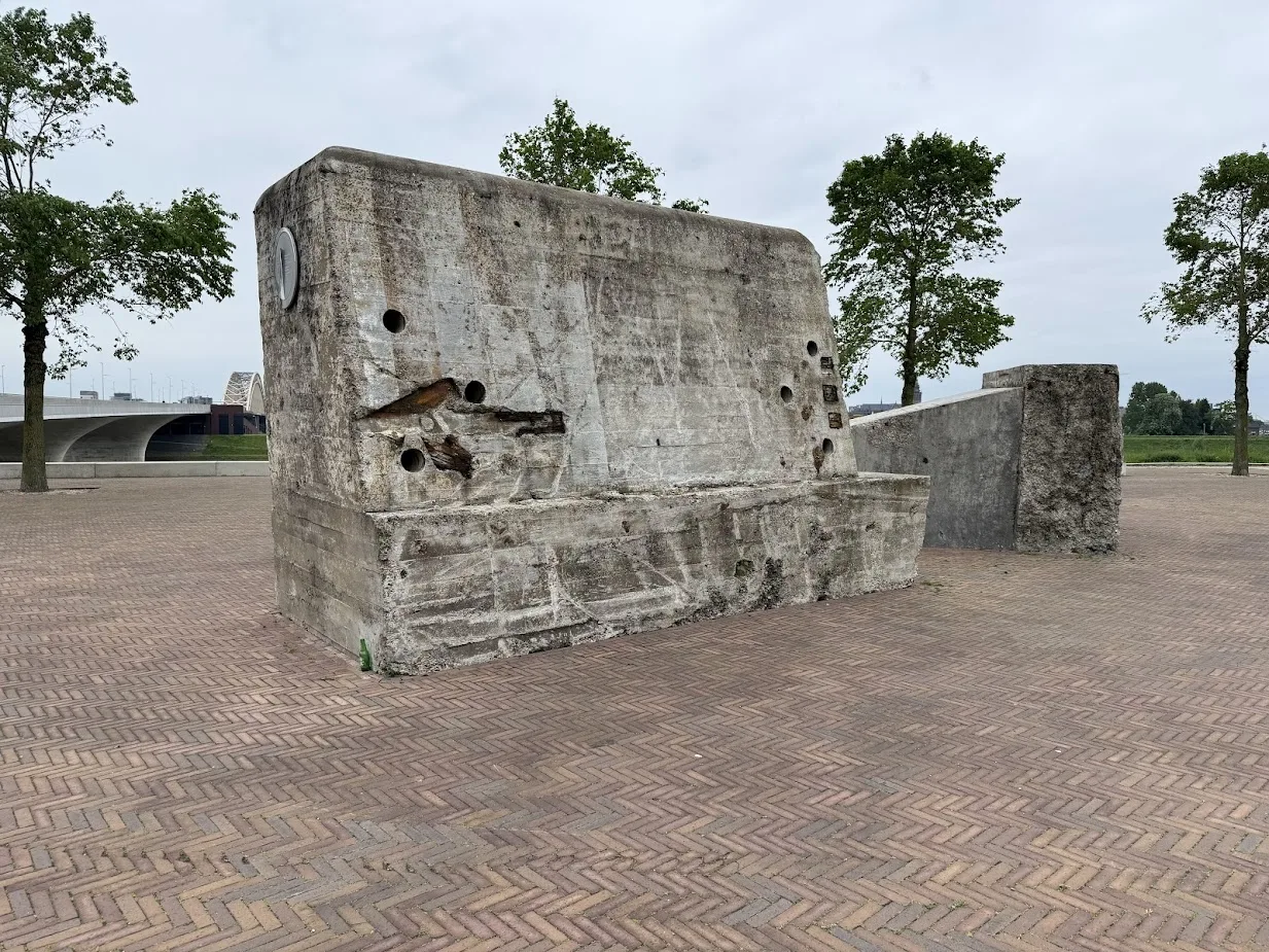Two weathered grey concrete anti-tank blocks standing on the north bank of the Waal at Nijmegen, one with traces of red-and-white paint.