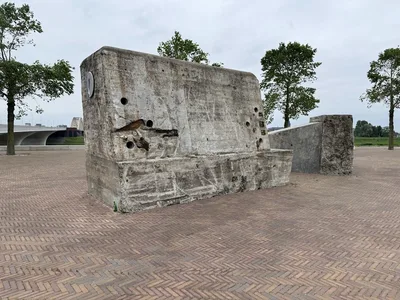 Two weathered grey concrete anti-tank blocks standing on the north bank of the Waal at Nijmegen, one with traces of red-and-white paint.