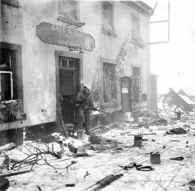 An American soldier with an M1 rifle outside the shell-damaged Café Bertemes in Marnach, abandoned German equipment lying in the snow.