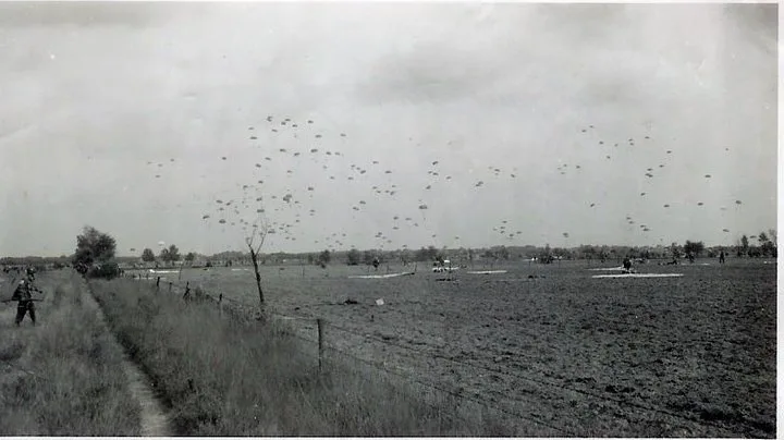 Black and white photo of dozens of parachutes coming down over flat fields near Eerde, with a fence and a sandy path in the foreground