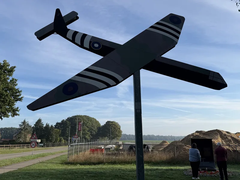 A model of a British glider in invasion stripes on a tall pole beside the road at Wolfheze, with the Glider Pilot Regiment memorial in the background.