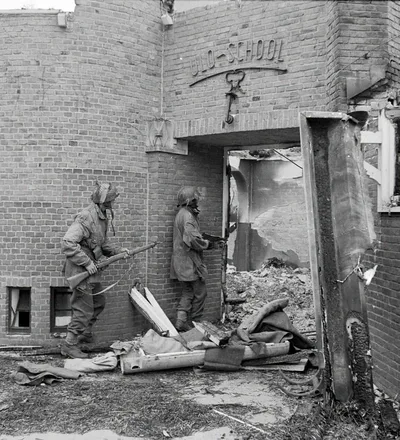 Two British glider pilots in helmets and Denison smocks, one with a rifle and one with a Sten gun, closing on the doorway of the burnt-out ULO school in Oosterbeek, September 1944.