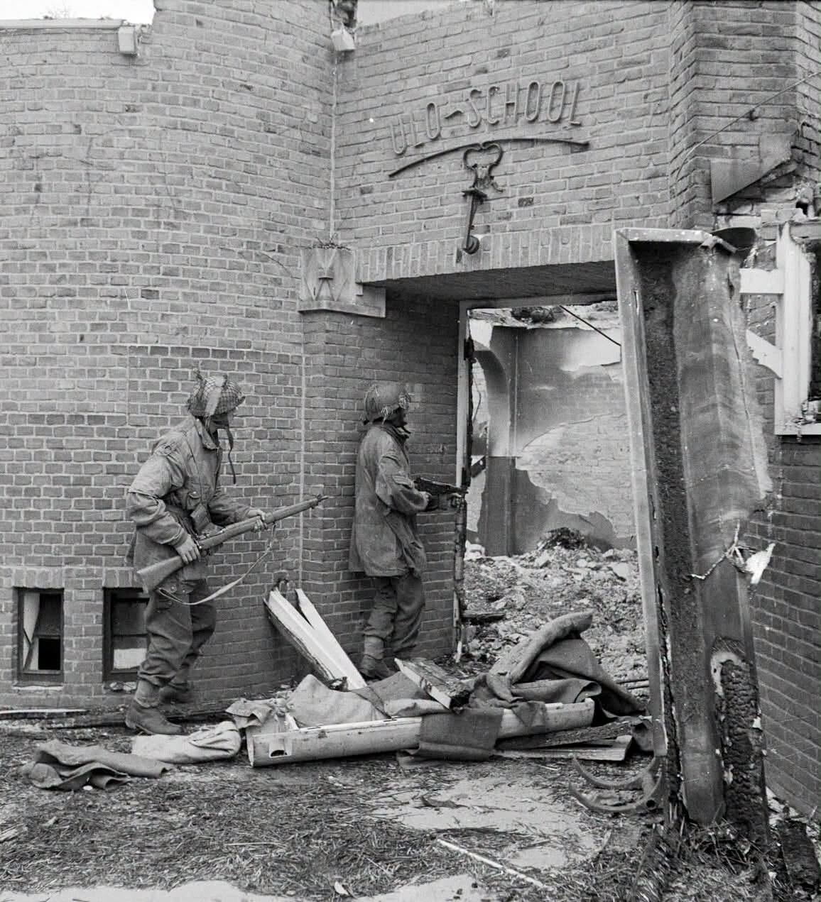 Two British glider pilots in helmets and Denison smocks, one with a rifle and one with a Sten gun, closing on the doorway of the burnt-out ULO school in Oosterbeek, September 1944.